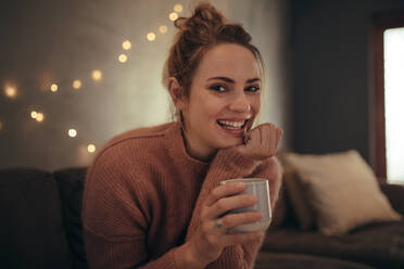 Schöne junge Frau sitzt zu Hause mit einer Tasse Kaffee. Lächelnde kaukasische Frau entspannt sich im Wohnzimmer und trinkt Kaffee. - JLPSF28283