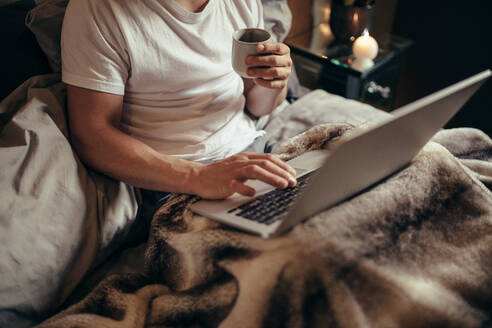 Cropped shot of man on bed using laptop and having coffee. Man working on laptop and drinking coffee on bed at home. - JLPSF28287