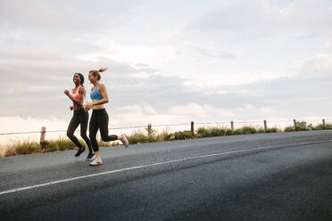 Zwei Frauen Athleten laufen auf der Straße früh am Morgen mit bewölktem Himmel im Hintergrund. Fitness Frauen joggen auf der Straße. - JLPSF28292