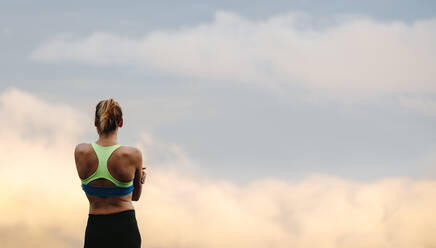 Rückansicht einer Sportlerin in Laufklamotten, die mit gefalteten Händen steht und in den offenen Himmel schaut, früh am Morgen. - JLPSF28293