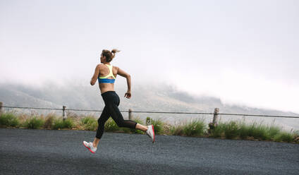 Woman athlete running on empty road early on a foggy morning with hills in the background. Fitness women sprinting on road. - JLPSF28294