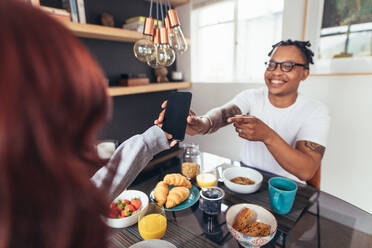 Afrikanischer Mann zeigt seiner Freundin am Frühstückstisch sitzend sein Mobiltelefon. - JLPSF28302
