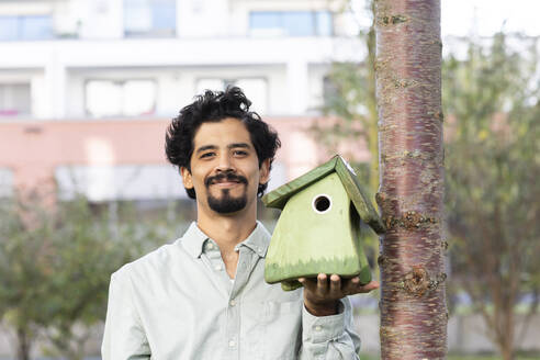 Lächelnder Mann hält grünes Vogelhaus am Baumstamm - SGF02925