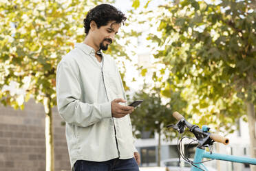 Smiling man using smart phone standing near bicycle - SGF02928