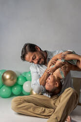 Vater spielt mit kleinem Baby, während er die Beine hält und es in einem mit Luftballons dekorierten Studio kitzelt, um gemeinsam Geburtstag zu feiern - ADSF40210