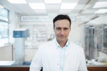 Mature pharmacist wearing lab coat standing in pharmacy - JOSEF14595