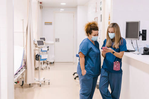 Ältere Krankenschwester im Gespräch mit einem Kollegen, der ein Smartphone in einer medizinischen Klinik benutzt - MMPF00475