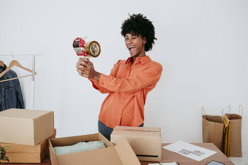 Cheerful young businesswoman with tape dispenser standing by desk - EBBF06854