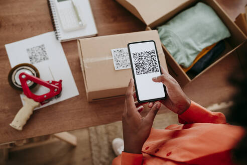 Hands of businesswoman scanning QR code through smart phone on package at desk - EBBF06856