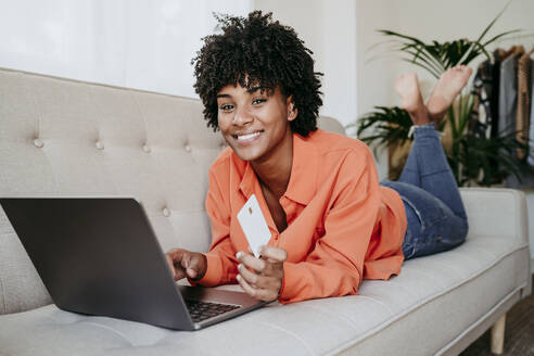 Happy young freelancer lying on sofa with laptop and credit card at home office - EBBF06906