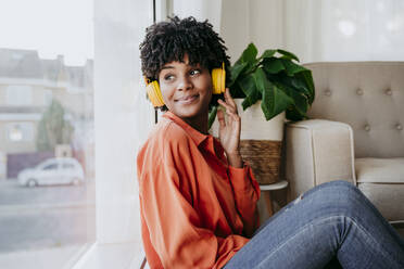 Smiling young freelancer listening to music through wireless headphones by window - EBBF06913