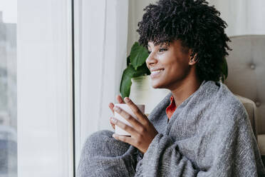Glückliche junge Frau mit Kaffeetasse, die zu Hause am Fenster sitzt - EBBF06921