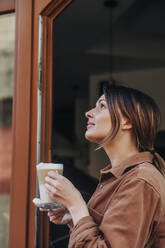 Thoughtful young woman with glass of cappuccino standing in doorway at cafe - VSNF00084