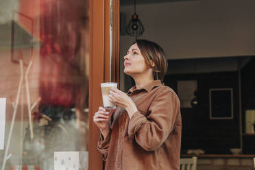 Nachdenkliche Frau mit einem Glas Cappuccino in der Tür eines Cafés stehend - VSNF00085