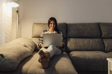 Happy woman using laptop on sofa in living room at home - EGHF00616