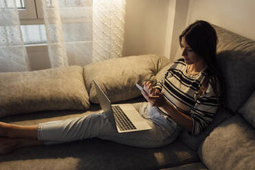 Frau sitzt mit Laptop und Smartphone auf dem Sofa zu Hause - EGHF00617