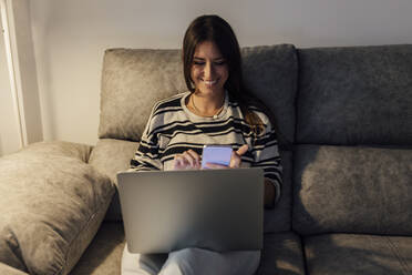 Glückliche Frau sitzt mit Laptop und Smartphone auf dem Sofa zu Hause - EGHF00618