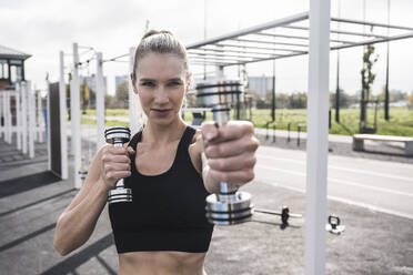 Smiling sportswoman exercising with dumbbells on sunny day - UUF27727