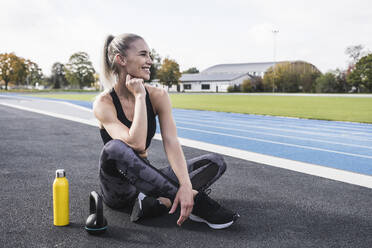 Lächelnde Sportlerin mit Kettlebell und Wasserflasche an der Strecke sitzend - UUF27739