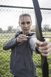 Young sportswoman doing archery in front of fence - UUF27807