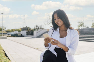 Happy young woman using smart phone sitting outdoors on sunny day - MEUF08332