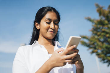 Lächelnde Frau Textnachrichten über Handy unter blauem Himmel - MEUF08341