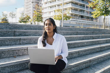 Thoughtful young woman sitting with laptop on steps - MEUF08347