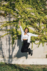 Happy young woman jumping in front of wall - MEUF08363