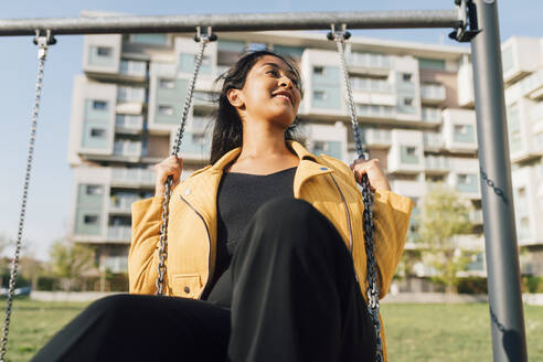 Playful young woman sitting on swing at playground - MEUF08372