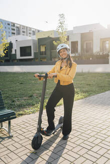 Glückliche junge Frau mit Elektro-Scooter an einem sonnigen Tag - MEUF08388