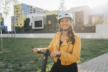 Lächelnde junge Frau mit Helm, die mit einem Tretroller im Park an einem sonnigen Tag steht - MEUF08389