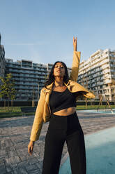 Young woman with arm raised standing in front of buildings at sunset - MEUF08413