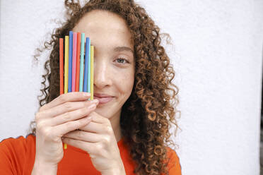 Smiling woman covering eye with colored pencils in front of white wall - AMWF00982