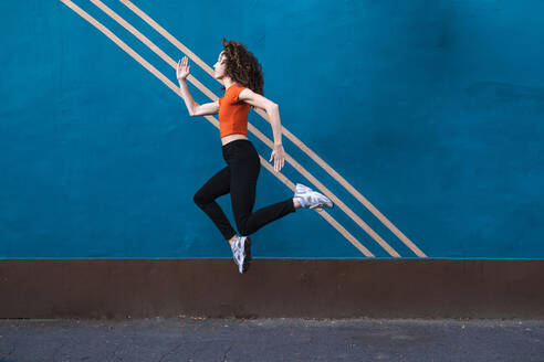 Junge Frau springt auf dem Fußweg vor der blauen Wand - AMWF00987