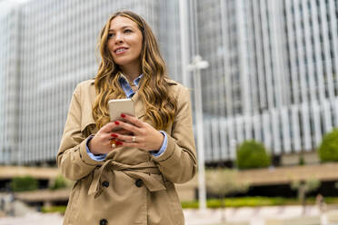 Lächelnde Geschäftsfrau mit Handy in der Hand vor einem Bürogebäude - DLTSF03367