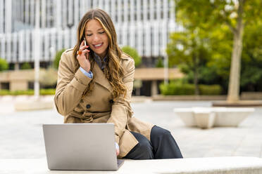 Lächelnde Geschäftsfrau, die mit einem Mobiltelefon spricht und einen Laptop im Büropark benutzt - DLTSF03370