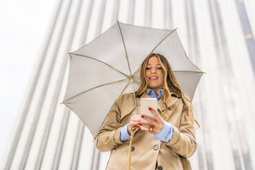Lächelnde Geschäftsfrau mit Regenschirm und Smartphone vor einem Gebäude - DLTSF03374