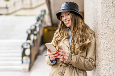 Smiling businesswoman wearing hat using mobile phone leaning on wall - DLTSF03379