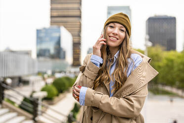 Smiling businesswoman wearing knit hat talking on mobile phone in front of buildings - DLTSF03389