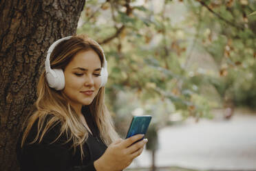 Lächelnde Frau, die über drahtlose Kopfhörer Musik hört und ihr Smartphone im Park benutzt - JBUF00120