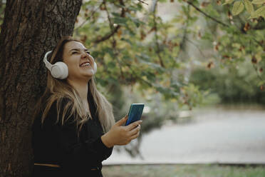 Glückliche Frau hört Musik über kabellose Kopfhörer und hält ihr Smartphone im Park - JBUF00121