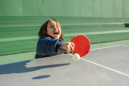 Fröhlicher Junge spielt Tischtennis an einem sonnigen Tag - ANAF00469