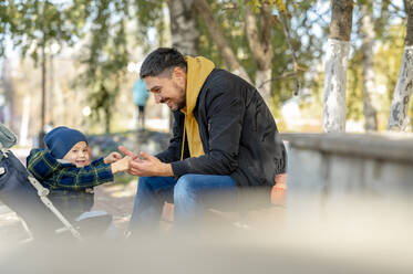 Glücklicher kleiner Junge im Kinderwagen, der die Hände des Vaters hält, der unter einem Baum im Park sitzt - ANAF00479