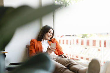 Smiling businesswoman holding mobile phone looking through window in office - JOSEF14645