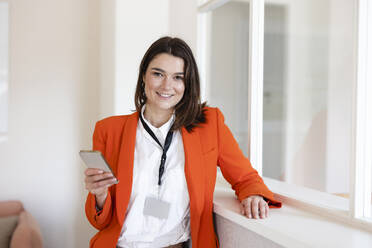 Happy businesswoman holding mobile phone in office - JOSEF14652