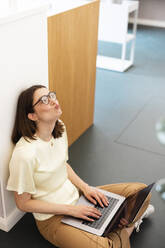 Gelangweilte Geschäftsfrau sitzt mit Laptop auf dem Boden im Büro - JOSEF14661