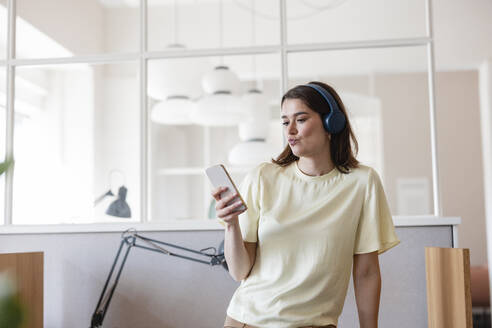 Geschäftsfrau hört Musik über drahtlose Kopfhörer und benutzt ein Mobiltelefon im Büro - JOSEF14669