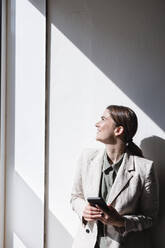 Happy businesswoman holding mobile phone leaning on wall - JOSEF14677