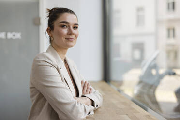 Confident businesswoman with arms crossed in front of window at office - JOSEF14698