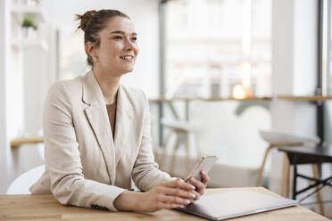 Smiling businesswoman holding mobile phone sitting with laptop at desk - JOSEF14710